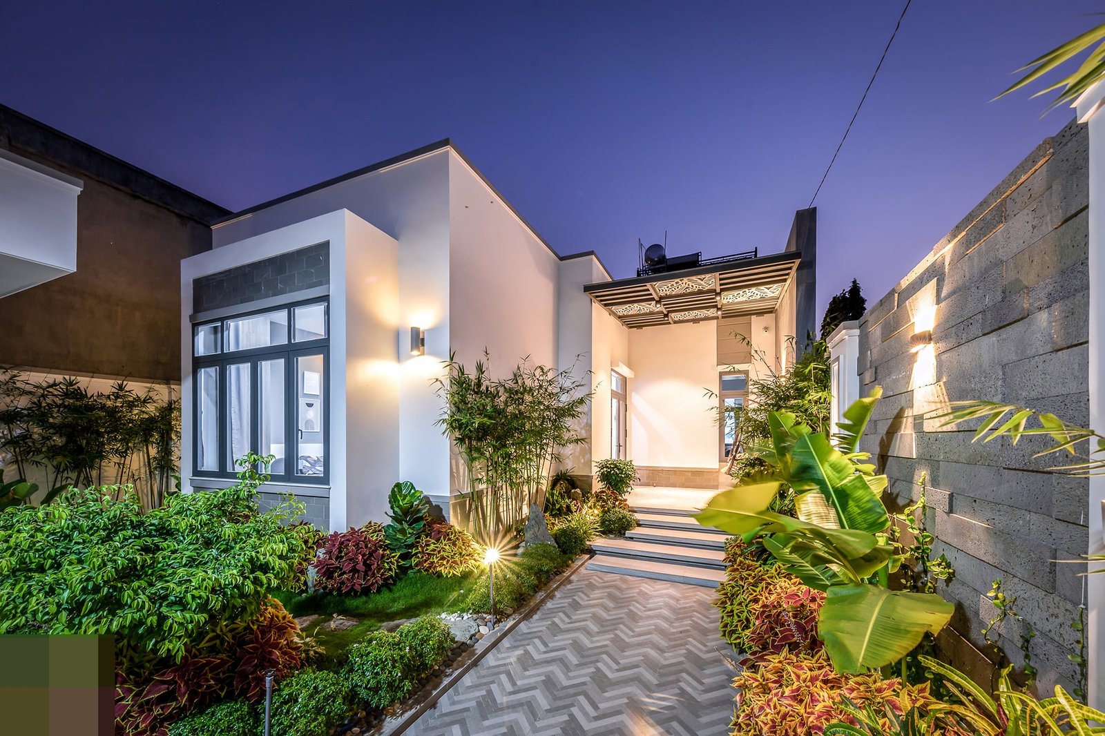 Nhà hiện đại có khu vườn xanh mát và lối đi lát gạch, ánh sáng đèn tạo cảm giác ấm áp vào buổi tối.